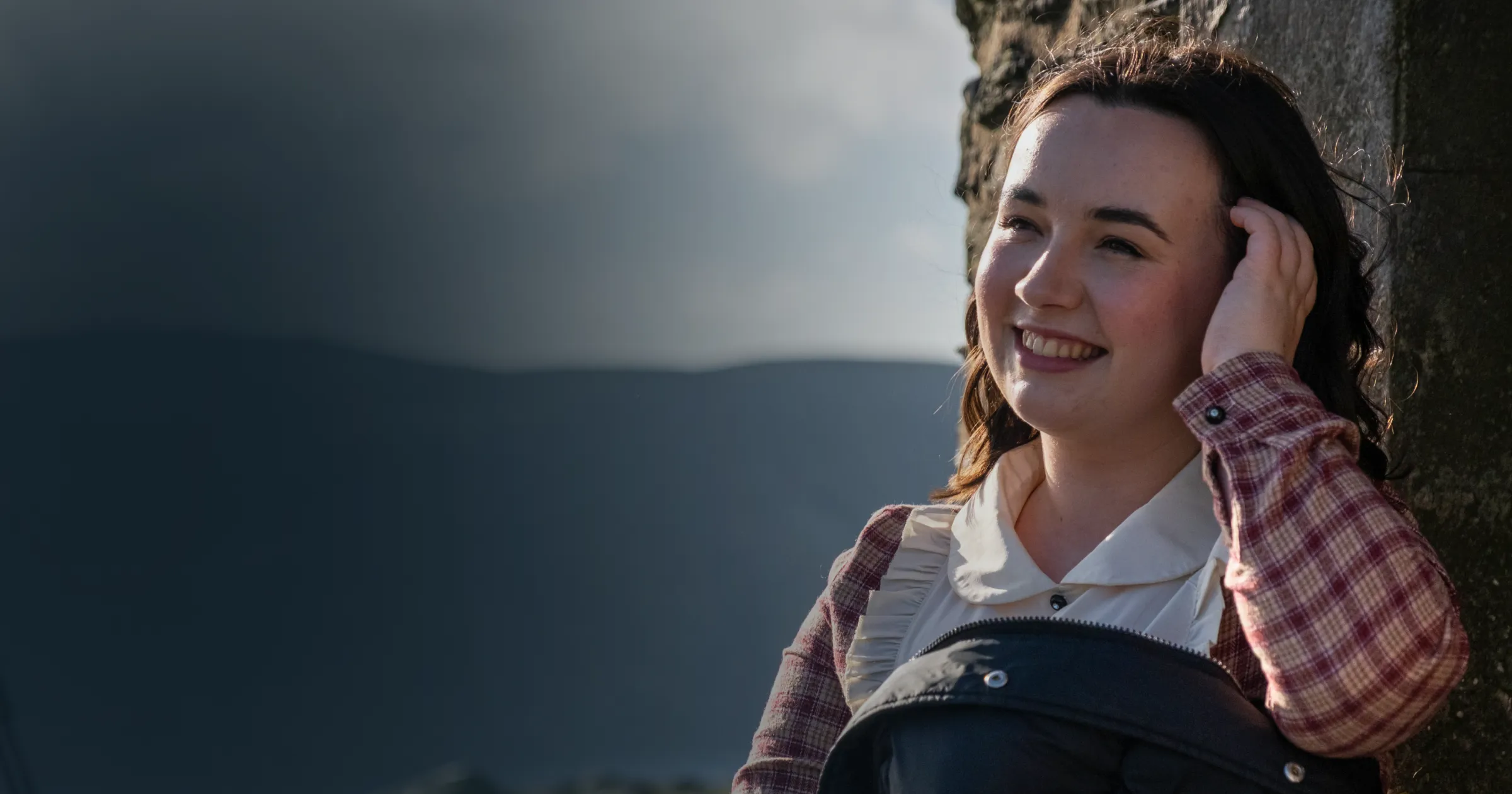 Smiling woman in a plaid shirt stands against a stone wall, hand in hair. Dark, moody sky and distant mountains create a contrast, adding drama.